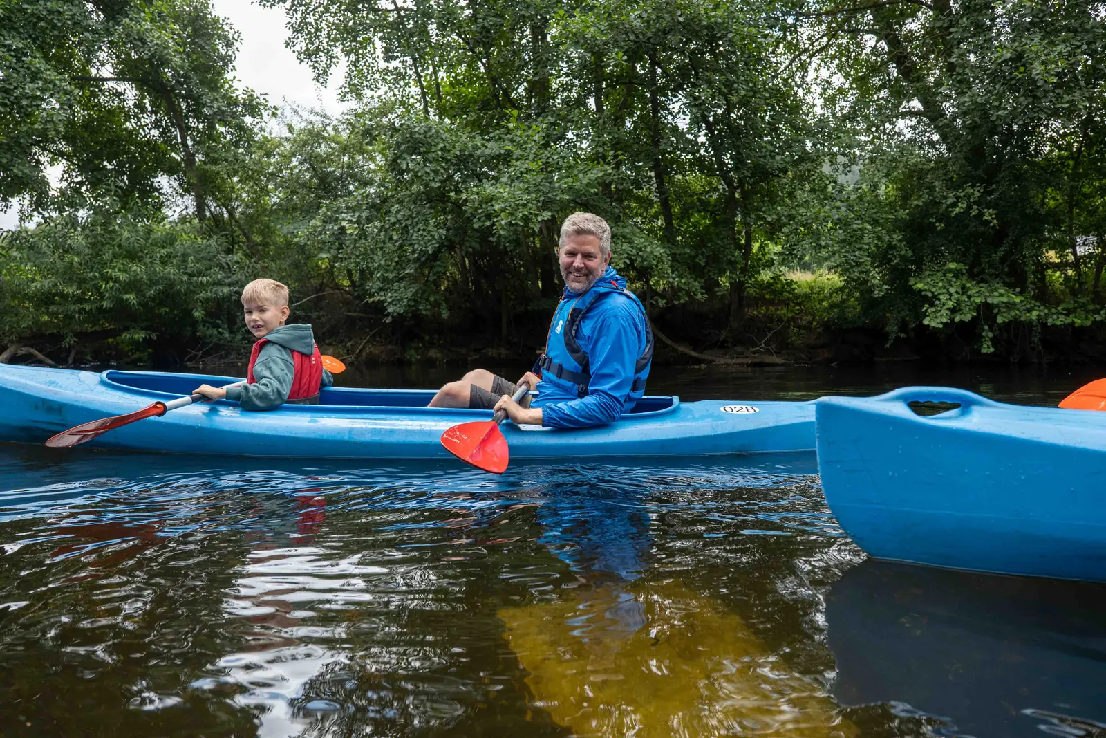 Vater und Sohn lachen im blauen Doppelkajak auf der Rur in die Kamera